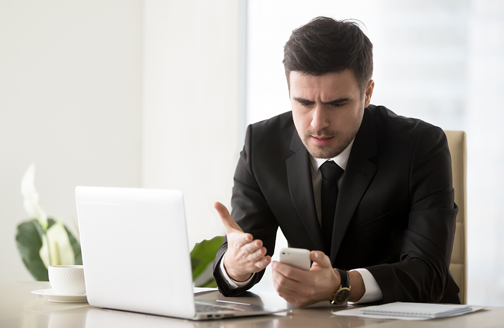 Angry businessman annoyed with phone call while working on laptop at office, mobile poor signal coverage